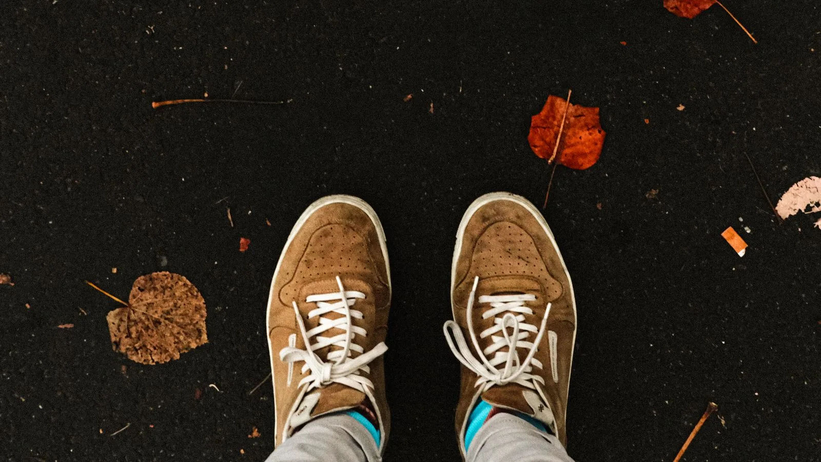 Brown sneakers on a dark pavement with scattered autumn leaves, embodying a fall vibe.